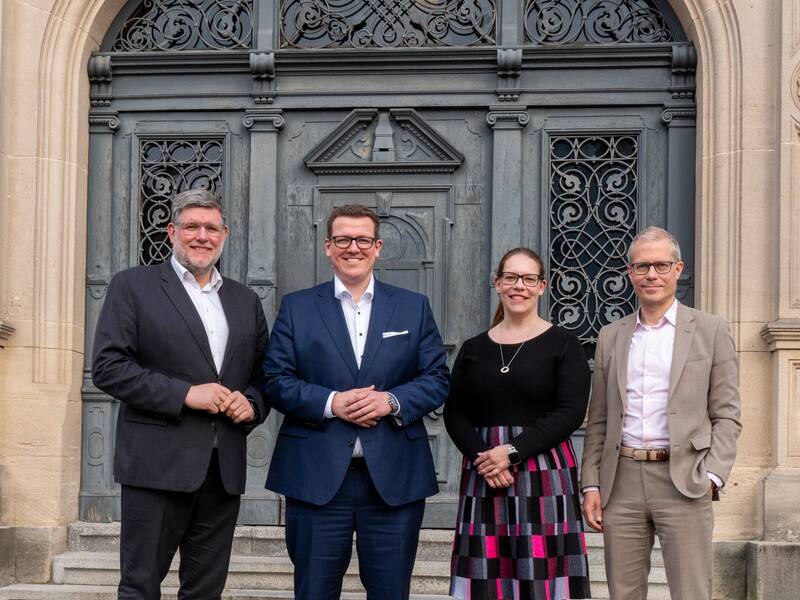 Vier Personen stehen in formeller Kleidung vor einem großen Holztor mit kunstvollen Verzierungen und einem Schild mit der Aufschrift "Landratsamt". Es handelt sich von links nach rechts um Landrat Dr. Ulrich Fiedler, Dominik Nusser. Dr. Julia Reuter und Prof. Dr. Boris Augurzky