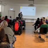 In einem hellen Raum mit großen Fenstern sitzt eine Gruppe von Menschen auf Stühlen und schaut zu Landrat Dr. Fiedler, der vor einer Leinwand steht, auf der eine Präsentation gezeigt wird.