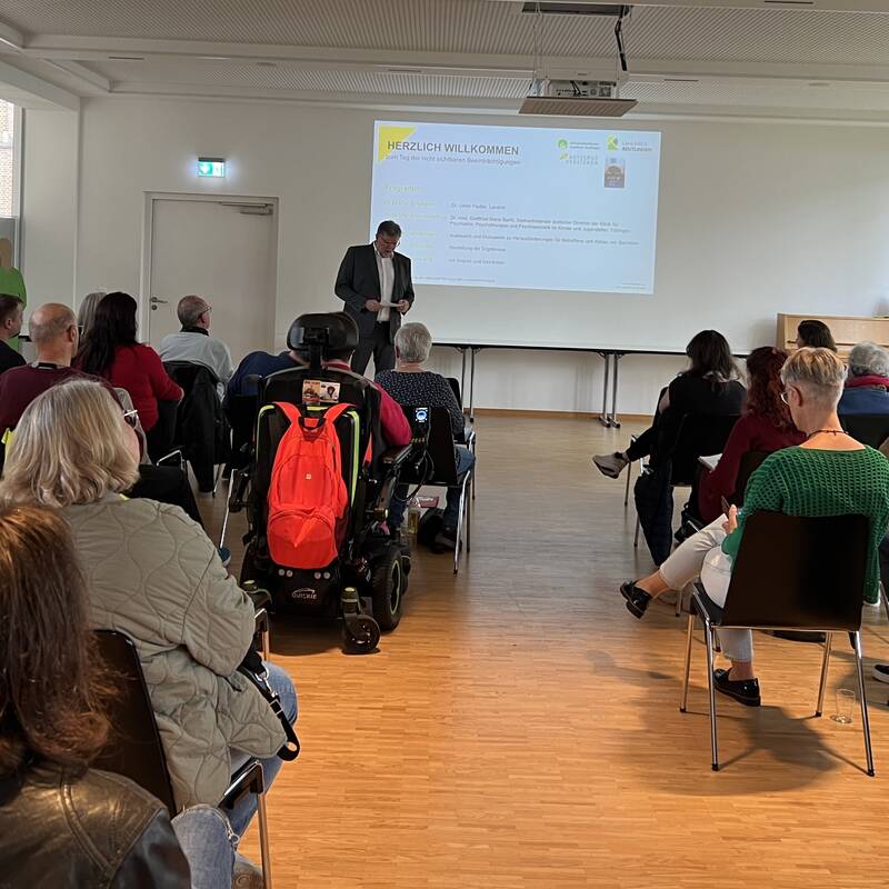 In einem hellen Raum mit großen Fenstern sitzt eine Gruppe von Menschen auf Stühlen und schaut zu Landrat Dr. Fiedler, der vor einer Leinwand steht, auf der eine Präsentation gezeigt wird.