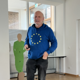 Dr. Gottfried Maria Barth steht in einem Raum vor einem Pappaufsteller und hält einen Vortrag.