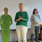 Drei Personen stehen in einem Raum, eine Frau hält einen Stift, während neben ihnen eine grüne Silhouette mit handschriftlichen Notizen zu sehen ist.