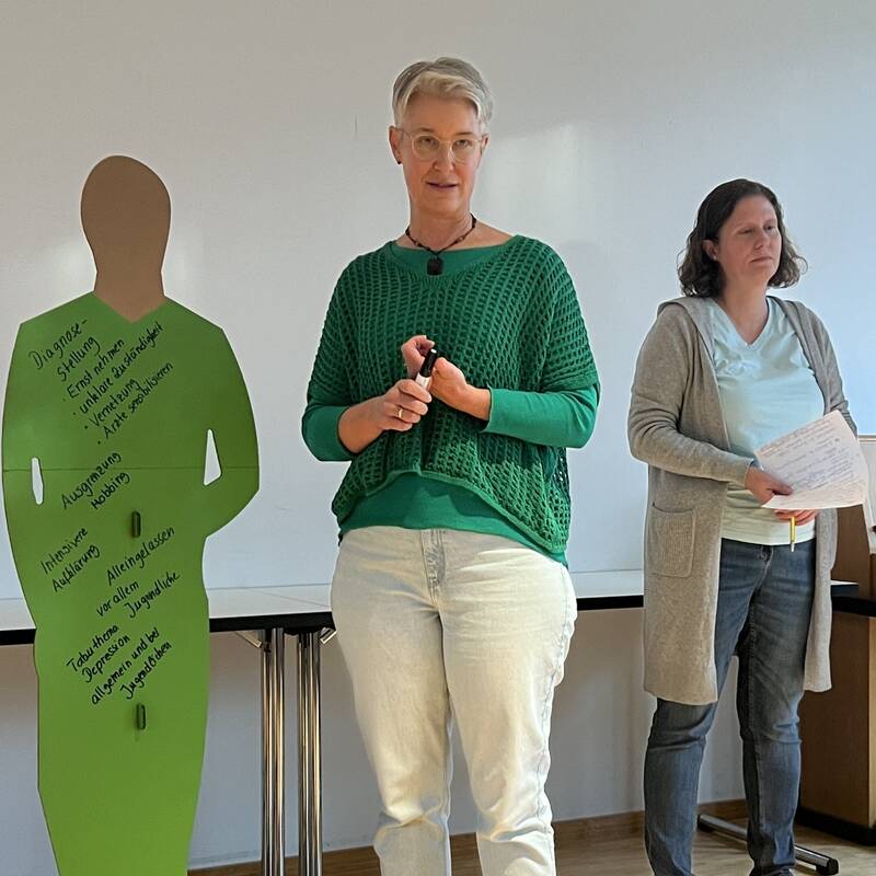 Drei Personen stehen in einem Raum, eine Frau hält einen Stift, während neben ihnen eine grüne Silhouette mit handschriftlichen Notizen zu sehen ist.