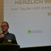 Eine Person spricht in ein Mikrofon vor einer Leinwand mit der Aufschrift "HERZLICH WILLKOMMEN zum Tag der nicht sichtbaren Beeinträchtigung 2025".