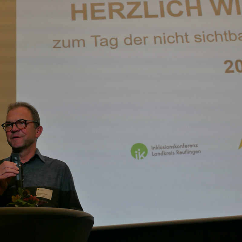 Eine Person spricht in ein Mikrofon vor einer Leinwand mit der Aufschrift "HERZLICH WILLKOMMEN zum Tag der nicht sichtbaren Beeinträchtigung 2025".