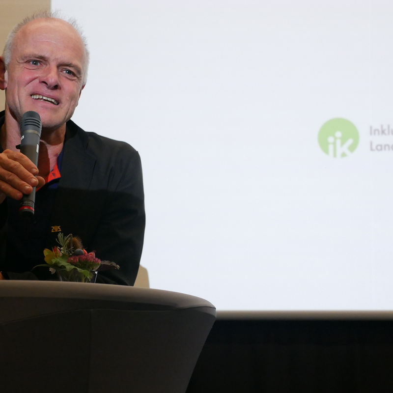Ein Mann steht an einem Tisch mit einem Mikrofon in der Hand und spricht vor einer Wand mit dem Logo der Inklusionskonferenz Landkreis Reutlingen.