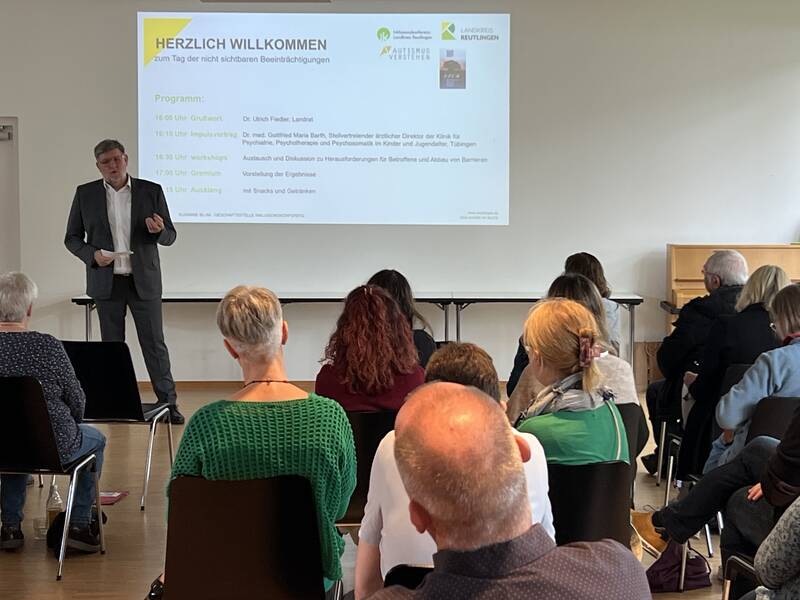 Landrat Dr. Ulrich Fiedler begrüßt eine Gruppe von sitzenden Zuhörern, während hinter ihm eine Leinwand mit Begrüßungstext und Programm abgebildet ist.