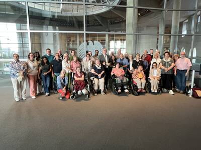Eine Gruppe von etwa 30 Menschen, darunter einige im Rollstuhl, posiert lächelnd vor einem modernen, gläsernen Innenraum.
