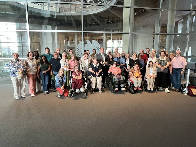 Eine Gruppe von etwa 30 Menschen, darunter einige im Rollstuhl, posiert lächelnd vor einem modernen, gläsernen Innenraum.