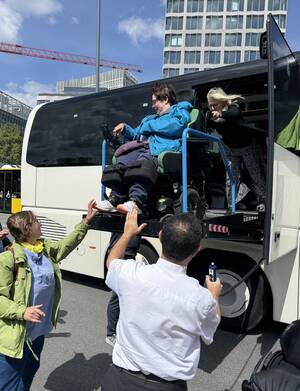 Eine Person in einem Rollstuhl wird mit einem Hebelift aus einem Bus herausgefahren, während mehrere Menschen dabei helfen und zuschauen.
