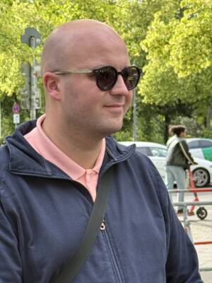 Ein Mann mit Sonnenbrille und blauer Jacke steht im Freien vor grünen Bäumen.