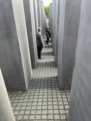 Zwei Personen stehen in einem schmalen gepflasterten Weg zwischen hohen, grauen Betonstelen, die Teil eines Denkmals sind.