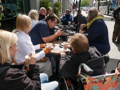 Eine Gruppe von Menschen sitzt an einem runden Tisch im Freien und genießt Getränke, darunter eine Person im Rollstuhl.