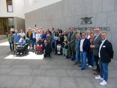 Eine Gruppe von Menschen, darunter einige im Rollstuhl, posiert lächelnd vor dem Gebäude der Landesvertretung Baden-Württemberg.