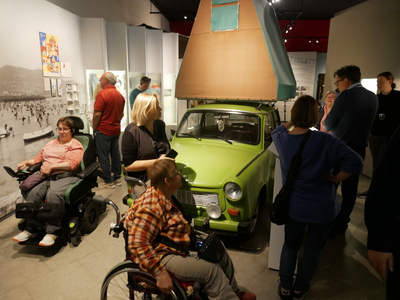 Mehrere Menschen, darunter Personen im Rollstuhl, betrachten in einem Ausstellungsraum ein grünes Auto mit einem aufgespannten Zeltdach.
