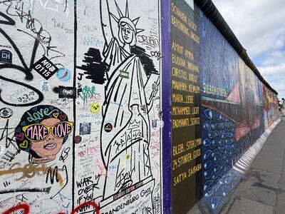 Das Bild zeigt ein mit Graffiti und bunten Kunstwerken bemaltes Stück der Berliner Mauer, darunter eine stilisierte Freiheitsstatue und verschiedene Schriftzüge.