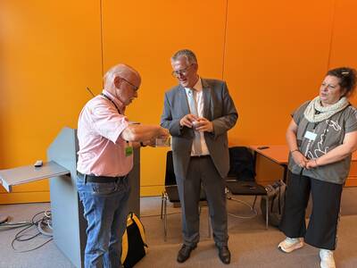 Zwei Männer und eine Frau stehen in einem Raum mit orangefarbener Wand. Ein Mann übergibt dem anderen Mann ein Geschenk,  während die Frau zuschaut.
