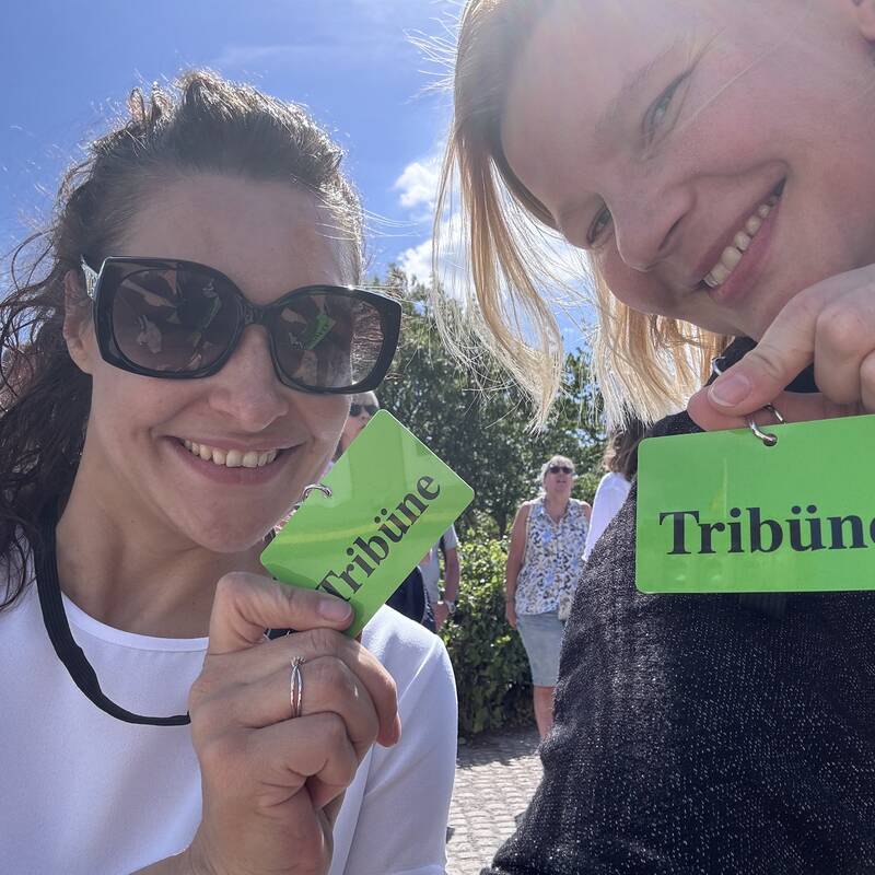 Zwei lächelnde Frauen halten grüne Schilder mit der Aufschrift „Tribüne“ in die Kamera.