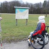 Eine Rollstuhlfahrerin und eine Frau mit Blinden-Langstock schauen auf eine Tafel mit Wanderwegen