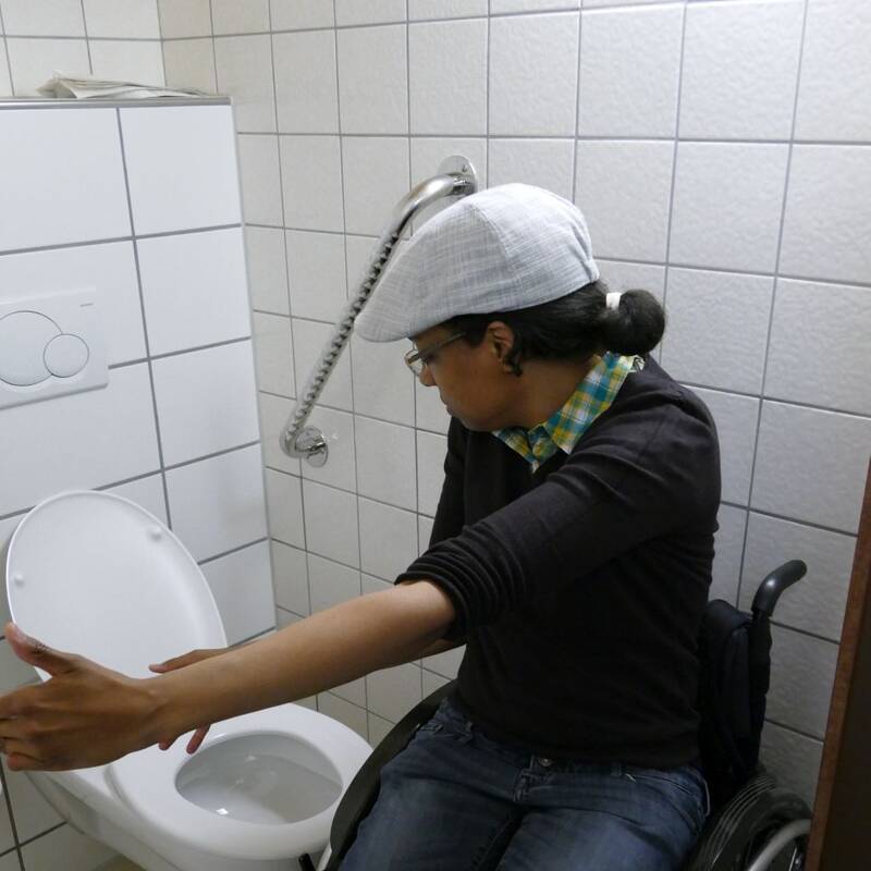 Eine Frau im Rollstuhl zeigt mit den Armen wieviel Platz im Behinderten-WC nötig ist. 