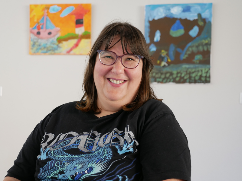Eine lächelnde Frau mit Brille und schwarzem T-Shirt sitzt vor zwei farbenfrohen abstrakten Gemälden an der Wand.
