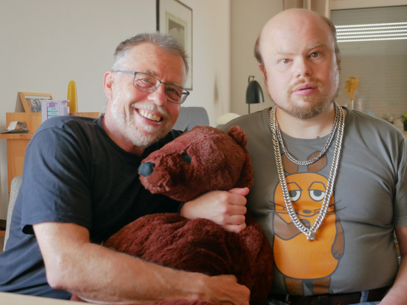 Zwei Männer sitzen nebeneinander in einer Wohnung, einer umarmt ein großes, braunes Stofftier, während der andere ein T-Shirt mit einem Comic-Motiv trägt.