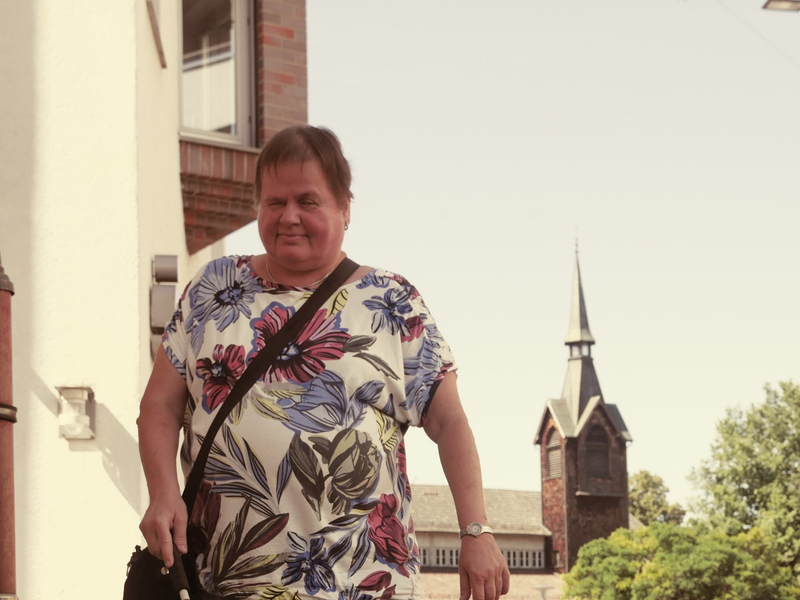 Eine Frau mit einem weißen Langstock geht auf einem Bürgersteig vor einem Backsteingebäude mit Turm entlang.