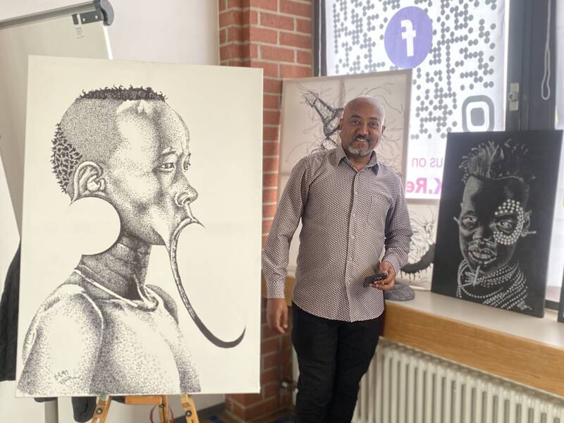 Ein Mann steht in einem Raum neben mehreren Kunstwerken, die Porträts zeigen, darunter ein großes Bild eines Profils mit verzierten Details auf einer Staffelei.