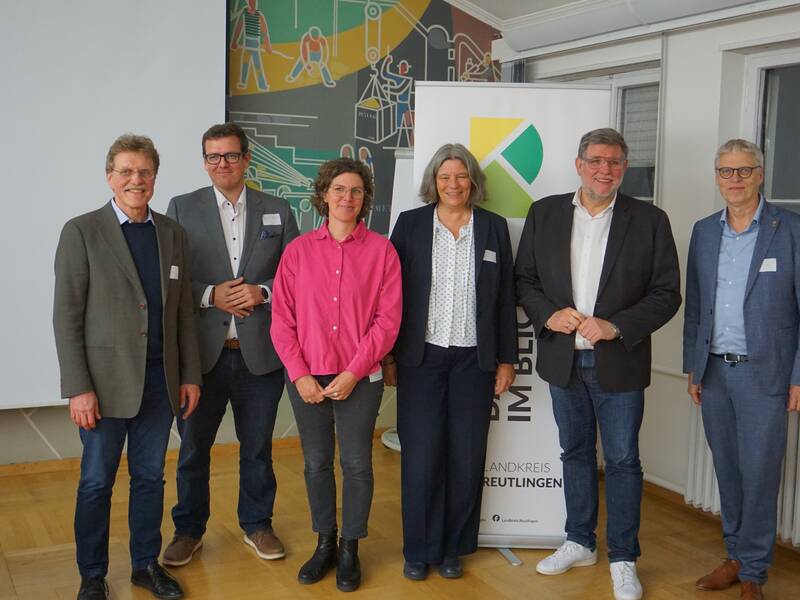 Sechs Personen posieren in einem Raum vor einem Banner mit einem Logo und einem bunten Wandbild im Hintergrund.