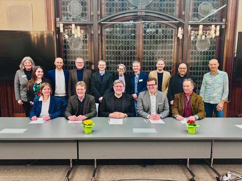 Eine Gruppe von fünfzehn Personen posiert lächelnd hinter einem Konferenztisch mit Blumenschmuck, vor einer verzierten Holz- und Glaskulisse.