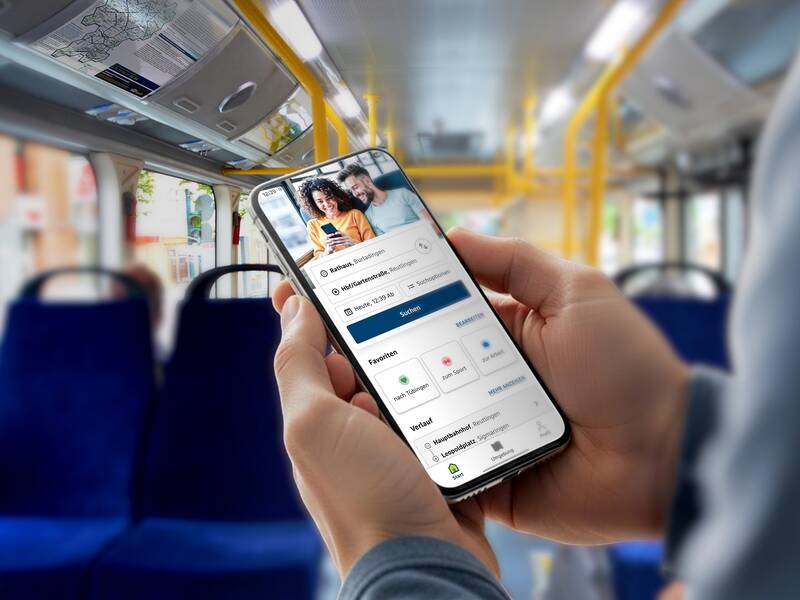 Eine Person hält ein Smartphone mit einer Reise-App in einem leeren Bus in der Hand.
