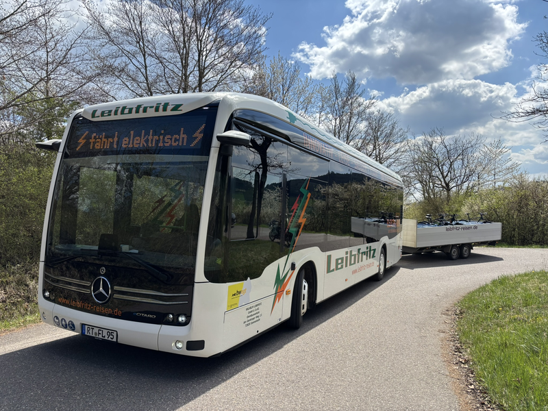 Ein weißer Elektrobus mit der Aufschrift "Leibfritz" zieht einen Anhänger durch eine landschaftliche Umgebung auf einer Straße.