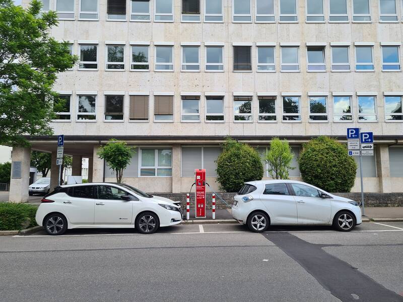 Zwei weiße Elektroautos parken nebeneinander vor einem Gebäude, dazwischen befindet sich eine Ladestation.