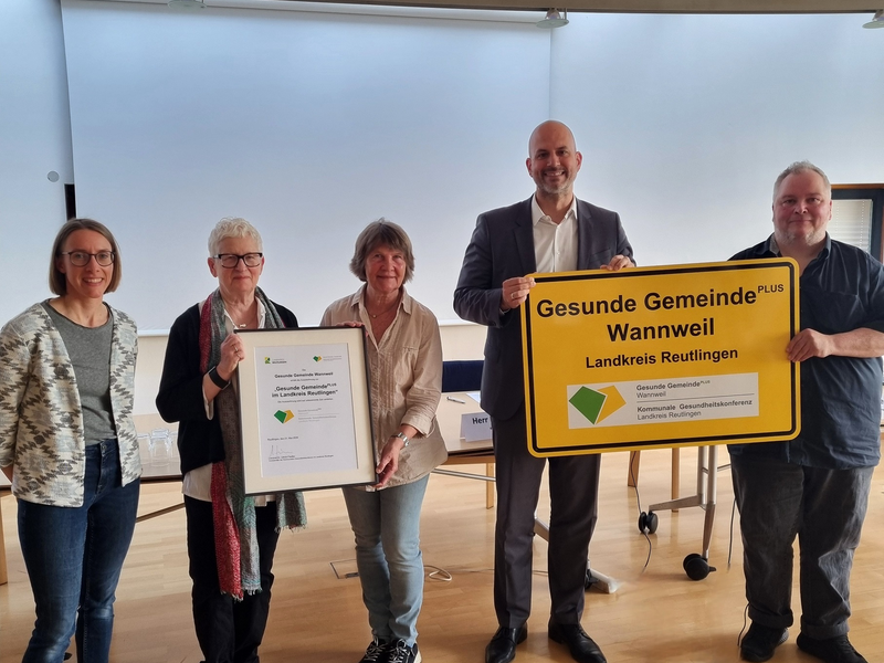 Fünf Personen stehen nebeneinander in einem Raum, wobei drei Personen eine gerahmte Urkunde und zwei Personen ein gelbes Schild mit der Aufschrift "Gesunde Gemeinde PLUS Wannweil Landkreis Reutlingen" halten.