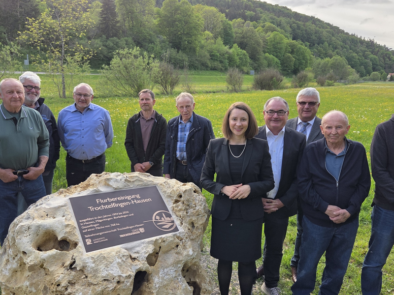 Eine Gruppe von Menschen steht auf einer Wiese hinter einem großen Stein mit einer Gedenktafel.