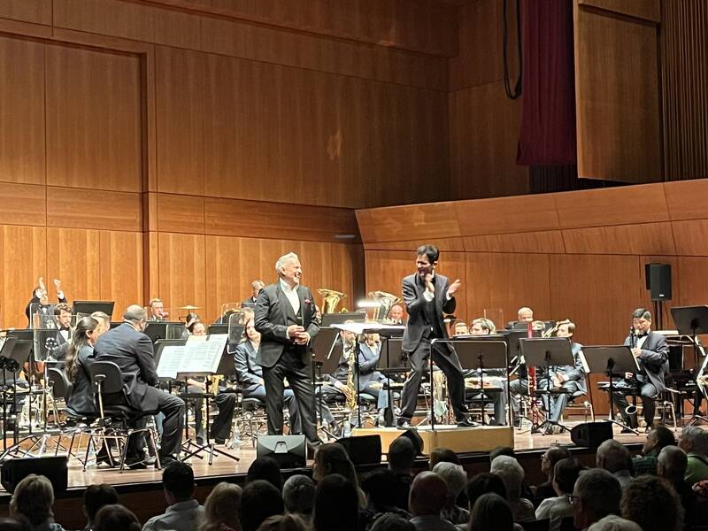 In einem Konzertsaal spielt ein Orchester auf der Bühne, während ein Moderator oder Dirigent das Publikum anspricht.