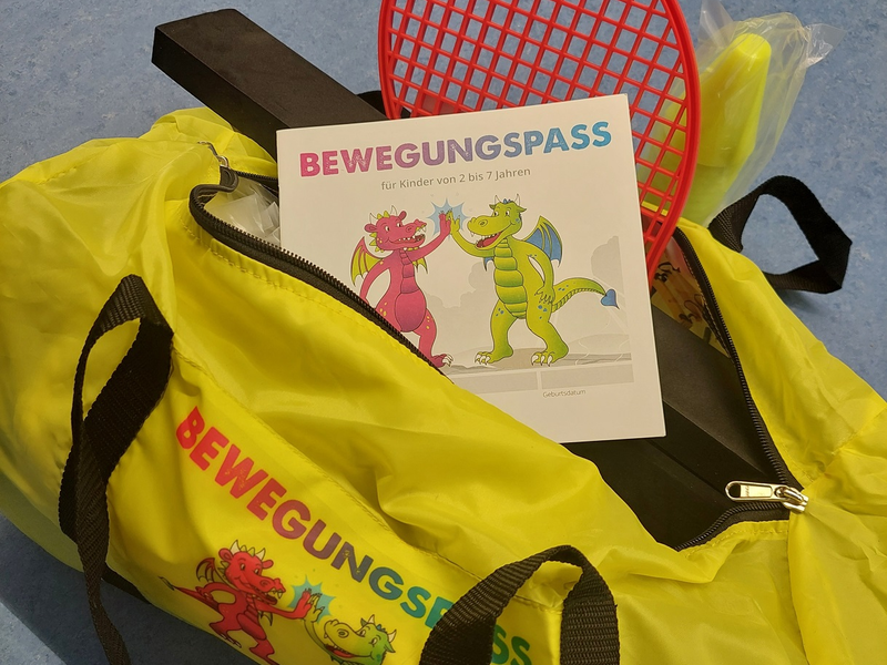 Eine gelbe Tasche mit der Aufschrift "BEWEGUNGSPASS" ist geöffnet und enthält einen roter Gitter-Schläger, ein Heft mit Drachenillustration und Zubehör für Kinderaktivitäten.