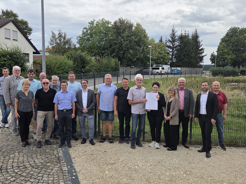 Eine Gruppe von Menschen unterschiedlichen Alters steht im Freien auf einem gepflasterten Weg vor einem Zaun und hält ein Dokument, umgeben von Bäumen und Wohnhäusern im Hintergrund.