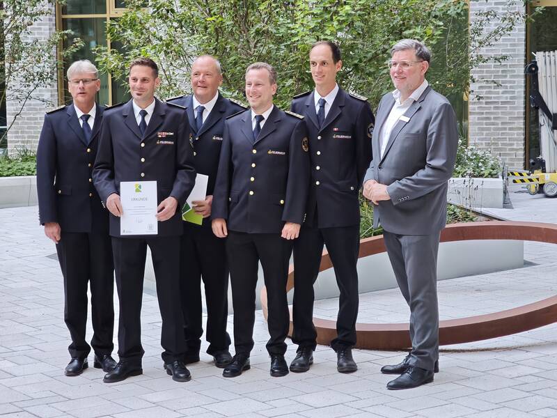 Sechs Männer, von denen fünf Feuerwehruniformen tragen und einer im Anzug, stehen zusammen und einer hält eine Urkunde in der Hand.