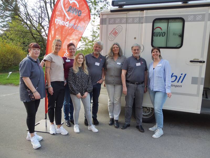 Eine Gruppe von acht Personen steht lächelnd vor einem medizinischen Mobil der AWO, neben einem orangefarbenen Banner.