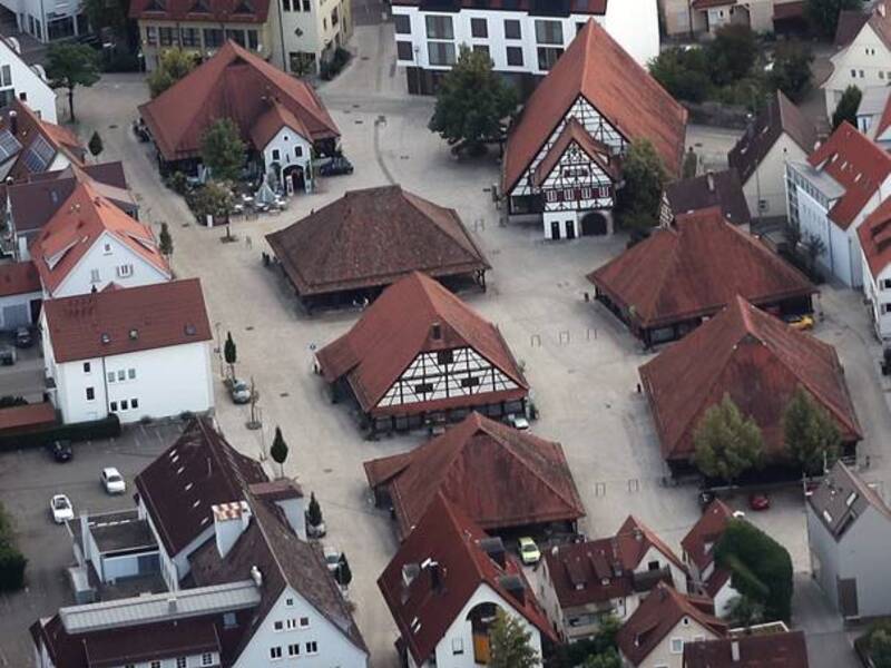 Luftaufnahme eines Dorfplatzes mit mehreren umgebenden Gebäuden im traditionellen Fachwerkstil und roten Dächern.