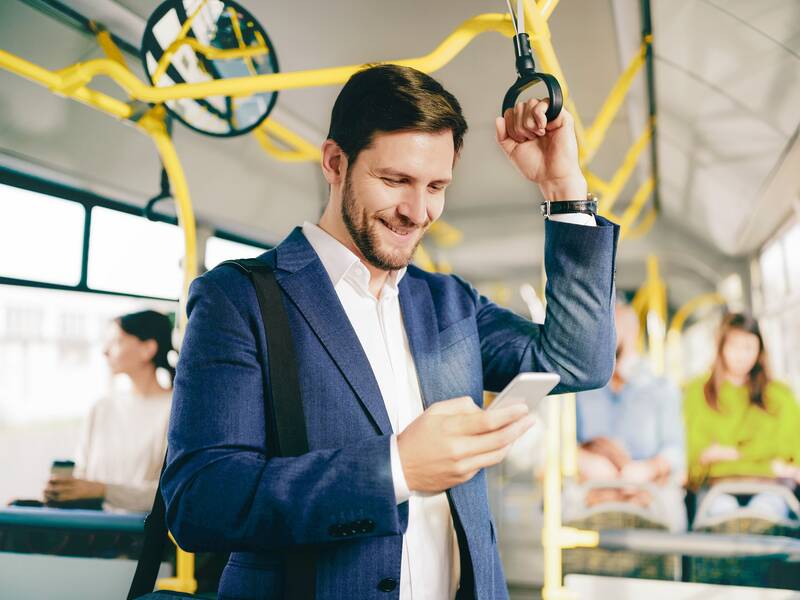 Ein Mann in einem Anzug steht in einem Bus und hält sich mit der linken Hand an einem Haltegriff fest. In der rechten Hand hält er ein Smartphone, auf das er schaut. Im Hintergrund sitzen weitere Passagiere im Bus.