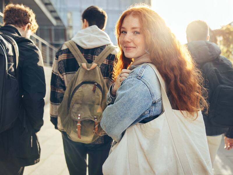 Eine junge Frau mit langen roten Haaren, die eine Umhängetasche trägt, dreht sich lächelnd auf einem sonnigen Gehweg zu mehreren Personen um.