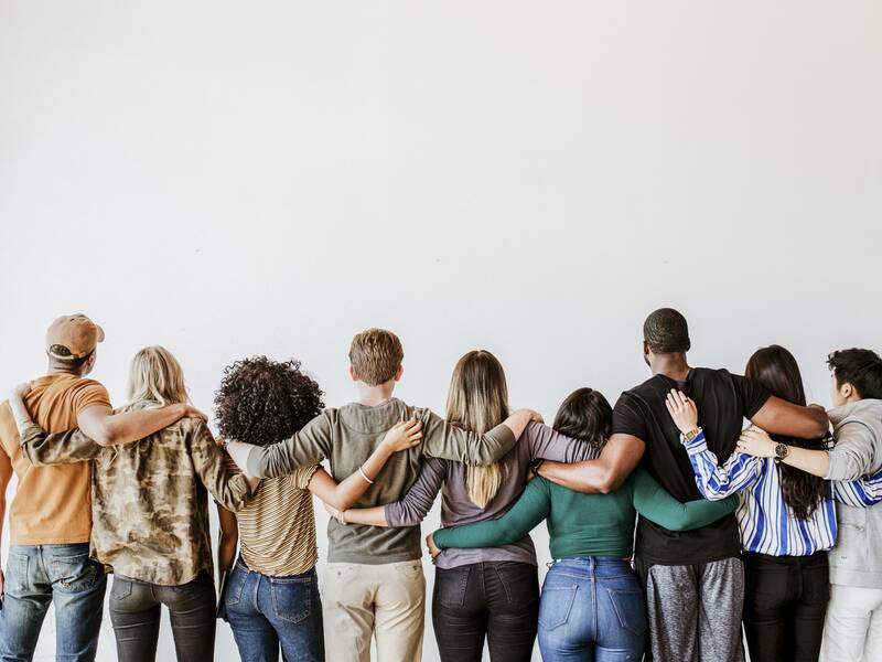 Eine Gruppe von acht Menschen steht Rücken an Rücken mit den Armen umeinander gelegt vor einer weißen Wand.
