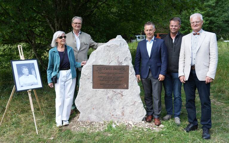 Eine Gruppe von fünf Personen steht neben einem Gedenkstein auf einer Wiese, mit einem gerahmten Schwarzweißfoto auf einem Ständer links von ihnen.