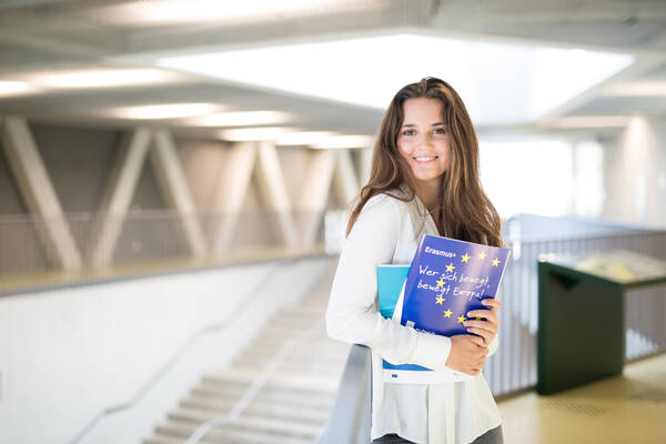 Eine lächelnde Person steht in einem modernen Gebäude und hält ein Buch mit der Aufschrift "Erasmus: Wer sich bewegt, bewegt Europa!" in der Hand.