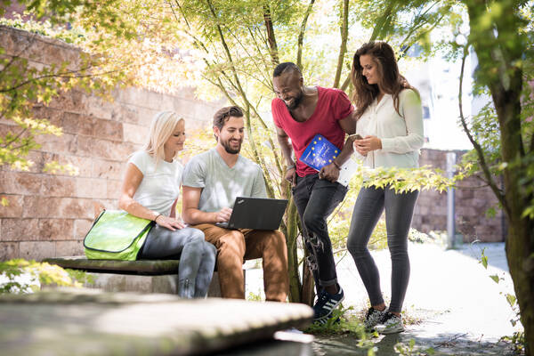 Vier junge Erwachsene, die im Freien nebeneinander stehen oder sitzen, schauen gemeinsam auf einen Laptop.
