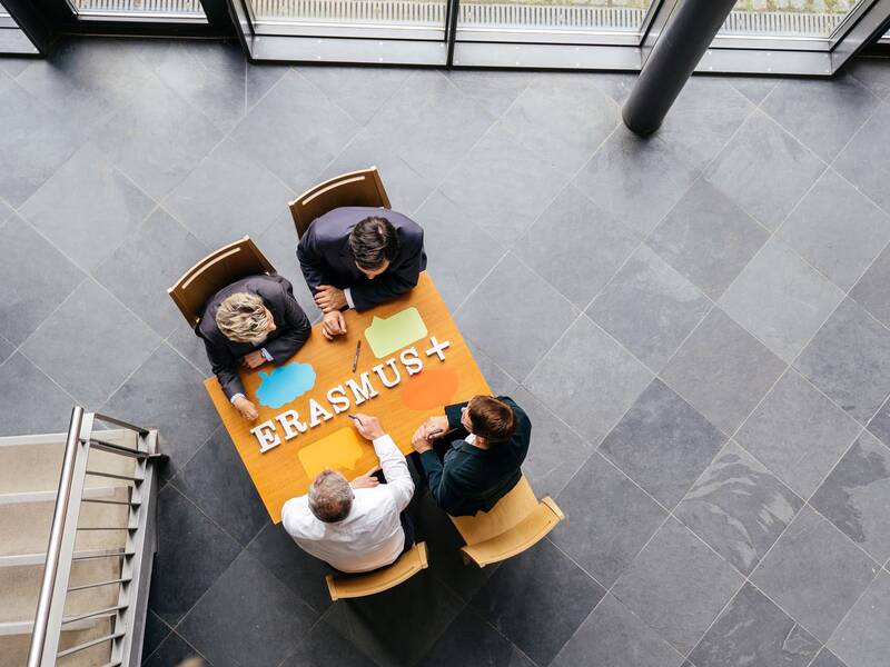 Vier Personen sitzen an einem Tisch, auf dem "ERASMUS+" steht, und scheinen ein Meeting abzuhalten.
