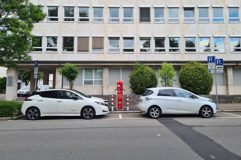 Zwei Elektro-Autos stehen an einer Ladesäule vor einem großen Gebäude.