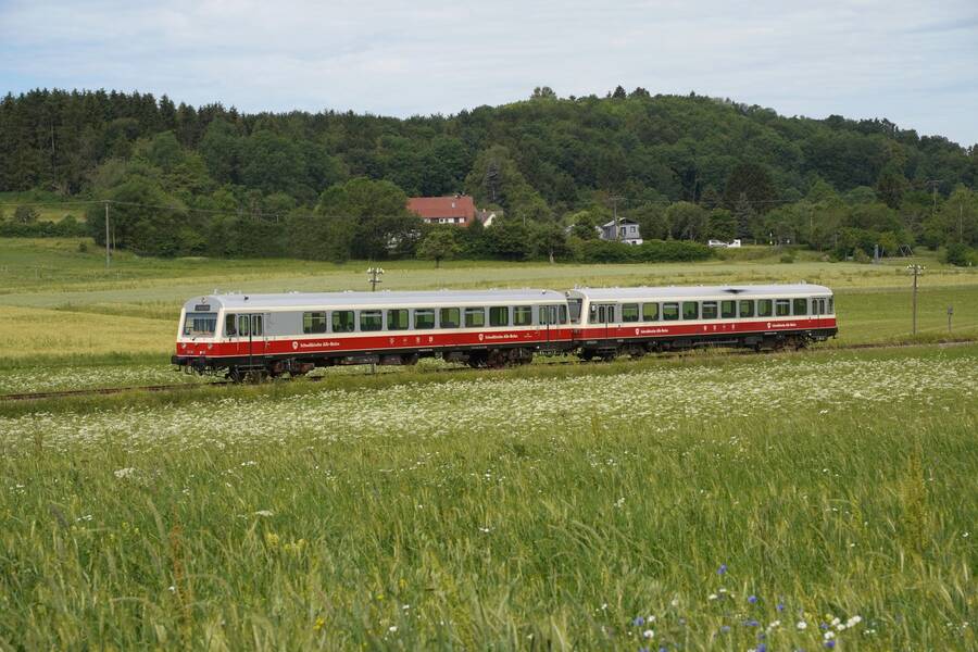 Ein roter Zug fährt durch eine grüne, ländliche Landschaft mit Wald und vereinzelten Häusern im Hintergrund.