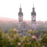 Im Bild sind zwei markante Kirchtürme einer barocken Kirche zu sehen, umgeben von sommerlicher Vegetation und im Hintergrund eine leicht nebelige Hügelkette.